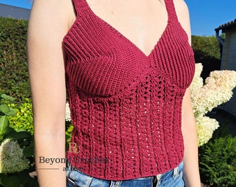 Puede incluir: Un top de crochet color granate con escote en V y un patrón de crochet. El top es sin mangas y tiene una longitud recortada. La modelo lleva el top con jeans azules.