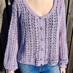 May include: A lilac crochet cardigan with a V-neck and long sleeves. The cardigan features five black and white buttons. The sleeves are gathered at the wrists. The person is wearing blue jeans. The cardigan is open-weave.