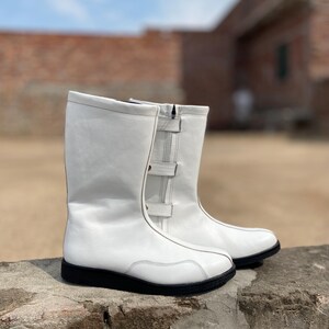 May include: A pair of white leather boots with black soles. The boots have a mid-calf height, a zipper, and two buckle straps on the side. The boots are sitting on a stone surface.
