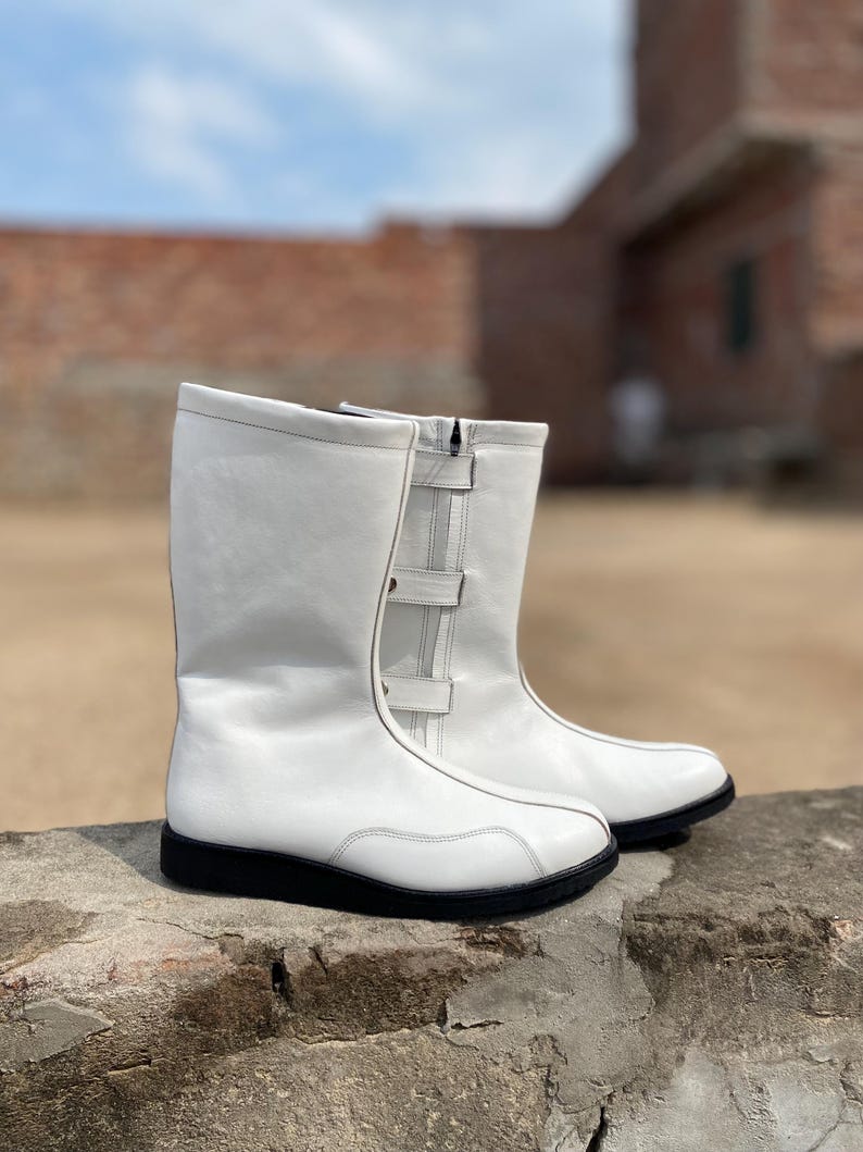 May include: A pair of white leather boots with black soles. The boots have a mid-calf height, a zipper, and two buckle straps on the side. The boots are sitting on a stone surface.