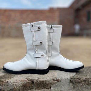 May include: A pair of white leather boots with black soles. The boots have a mid-calf height, with decorative straps and silver-tone hardware. The boots are set against a blurred background of a brick wall and a cloudy sky.