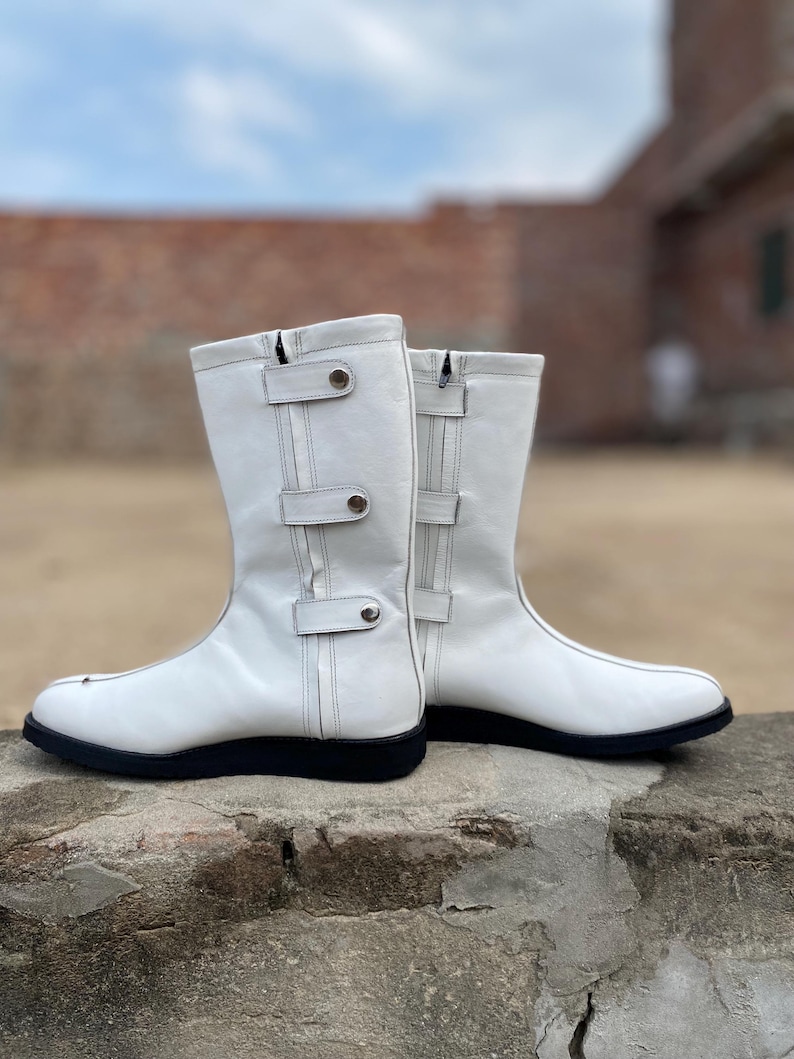 May include: A pair of white leather boots with black soles. The boots have a mid-calf height, with decorative straps and silver-tone hardware. The boots are set against a blurred background of a brick wall and a cloudy sky.