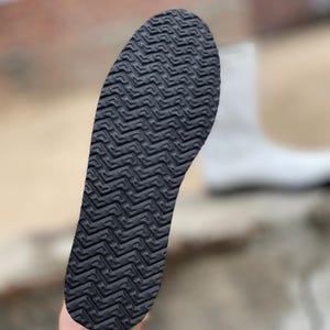 May include: Close-up of a black shoe sole with a textured, zig-zag pattern. The sole is held up against a blurred background, showcasing the intricate design and grip of the footwear. The sole is designed for traction.