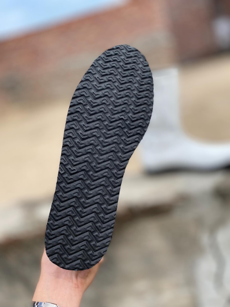 May include: Close-up of a black shoe sole with a textured, zig-zag pattern. The sole is held up against a blurred background, showcasing the intricate design and grip of the footwear. The sole is designed for traction.