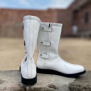 May include: A pair of white leather boots with black soles. The boots have a mid-calf height, a zipper, and decorative straps with silver-colored metal accents. The boots are set against a blurred outdoor background.