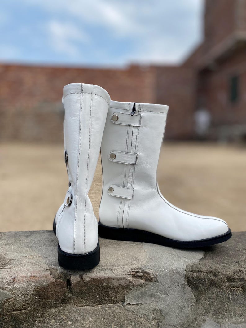 May include: A pair of white leather boots with black soles. The boots have a mid-calf height, a zipper, and decorative straps with silver-colored metal accents. The boots are set against a blurred outdoor background.