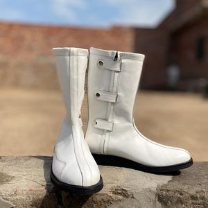 May include: A pair of white leather boots with black soles. The boots have a mid-calf height, a side zipper, and decorative straps with silver-colored buckles. The boots are set against a blurred outdoor background.