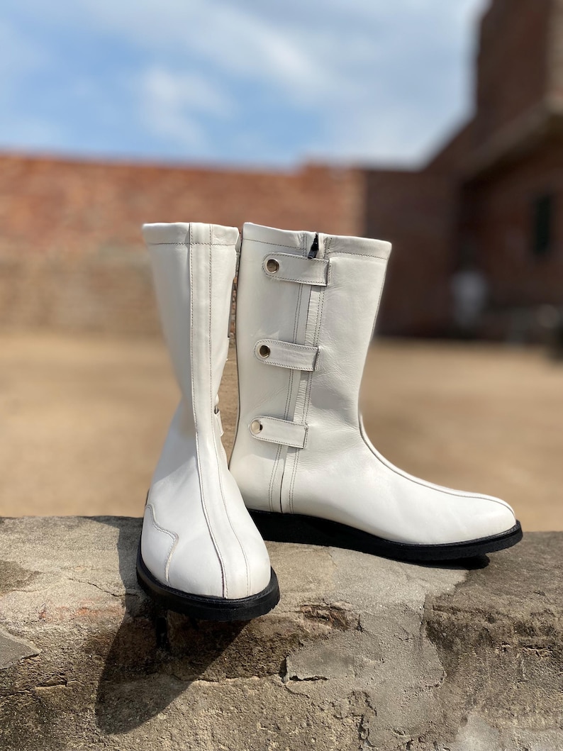 May include: A pair of white leather boots with black soles. The boots have a mid-calf height, a side zipper, and decorative straps with silver-colored buckles. The boots are set against a blurred outdoor background.