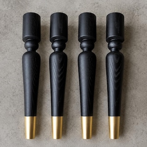May include: Four black wooden furniture legs with a gold-colored metal base. The legs have a turned design with a cylindrical top and a tapered body. The legs are displayed against a neutral gray background.