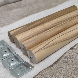 Puede incluir: Cuatro patas de muebles de madera con un acabado de vetas naturales, acompañadas de cuatro placas de montaje metálicas. Las patas son cilíndricas y parecen ser de madera dura. Las placas metálicas son cuadradas con agujeros para tornillos.