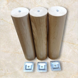 Può includere: Tre gambe per mobili in legno con finitura marrone chiaro, ciascuna di forma cilindrica e con parte superiore piatta. Le gambe sono alte circa 25 cm e sono accompagnate da tre piastre di montaggio in metallo.
