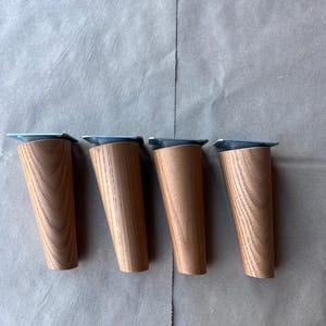 Könnte beinhalten: Ein Set aus vier konisch zulaufenden Möbelbeinen aus hellbraunem Holz mit sichtbarer Maserung. Jedes Bein verfügt über eine flache, silberfarbene Metallmontageplatte an der Oberseite, die für eine sichere Befestigung an Möbeln ausgelegt ist.