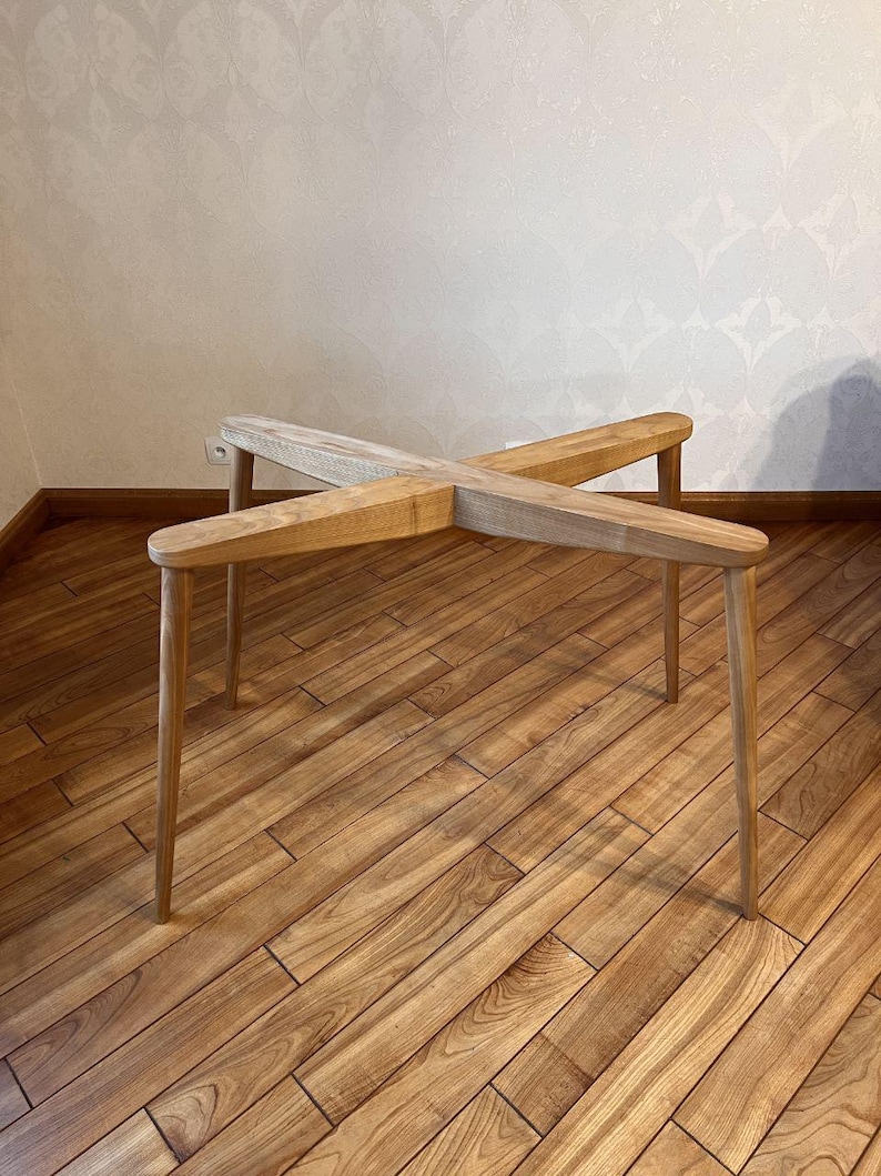May include: A light-colored wooden table with an X-shaped top and four slender legs. The table has a natural wood grain finish and a minimalist design. The table is approximately 24 inches long and 12 inches high.