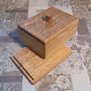 Op de afbeelding: Een rechthoekige houten botervloot met een deksel en een kleine knop. De botervloot staat op een bijpassende houten basis. De houtnerf is zichtbaar en de kleur is lichtbruin. De botervloot staat op een patroon oppervlak.