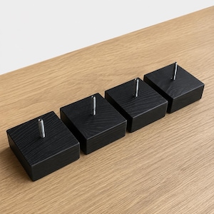 Puede incluir: Un juego de cuatro patas cuadradas para muebles hechas de madera negra con una textura de veta visible. Cada pieza cuenta con un perno roscado de metal centrado y vertical que sobresale de la superficie superior para su fijación.