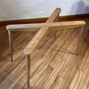 May include: A light-colored wooden table with a unique cross-shaped design. The table has four legs and a central support structure. The wood has a natural grain and a smooth finish. The table's dimensions are approximately 30 inches long and 30 inches wide.