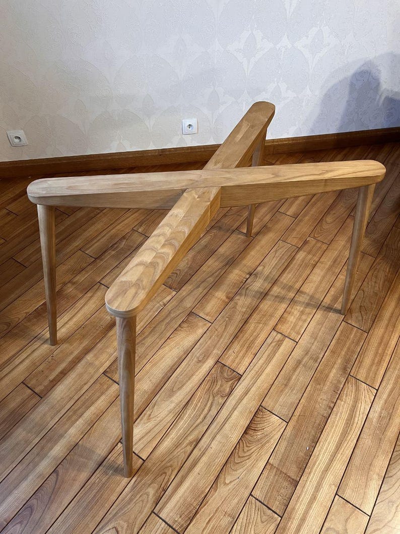 May include: A light-colored wooden table with a unique cross-shaped design. The table has four legs and a central support structure. The wood has a natural grain and a smooth finish. The table's dimensions are approximately 30 inches long and 30 inches wide.