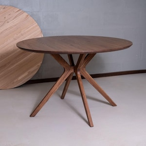 Puede incluir: Una mesa de comedor redonda de madera marrón oscuro con una superficie circular lisa y una base con cuatro patas anguladas que se unen en un punto central. La veta de la madera es visible y la mesa se apoya sobre un suelo de color claro.