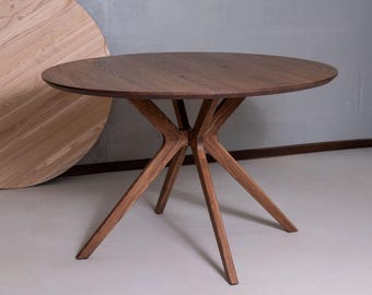 Puede incluir: Una mesa de comedor redonda de madera marrón oscuro con una superficie circular lisa y una base con cuatro patas anguladas que se unen en un punto central. La veta de la madera es visible y la mesa se apoya sobre un suelo de color claro.