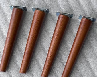 Puede incluir: Cuatro patas de mueble cónicas hechas de madera marrón con un patrón de veta visible. Cada pata cuenta con una placa de montaje de metal plateado en la parte superior, diseñada para fijarse a las bases de los muebles. Están alineadas.