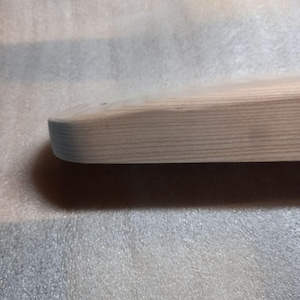 May include: Close-up of a light-colored, unfinished wooden board with rounded edges. The wood grain is visible, and the board appears smooth. The board is likely for crafting or woodworking projects.