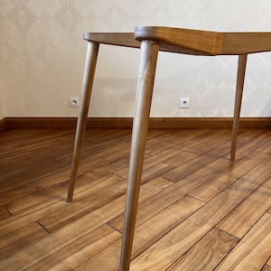 May include: A light-colored wooden table with four angled legs. The table's top is a light brown color, and the legs are a lighter shade of brown. The table is set on a wooden floor.