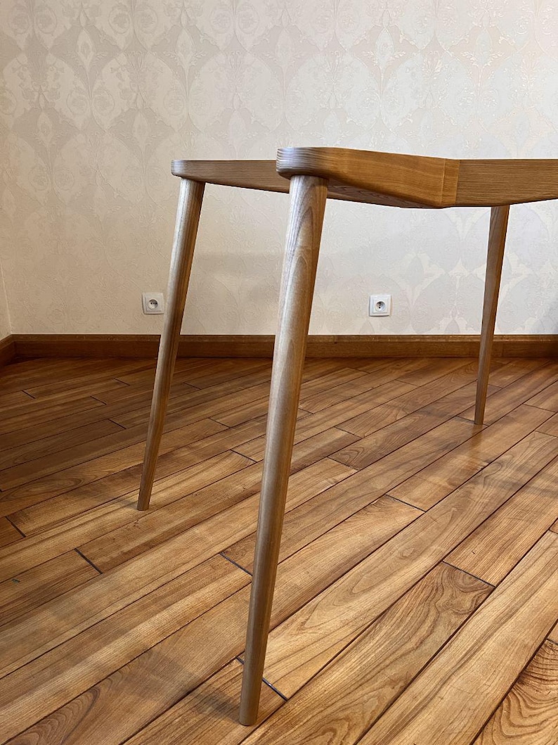 May include: A light-colored wooden table with four angled legs. The table's top is a light brown color, and the legs are a lighter shade of brown. The table is set on a wooden floor.