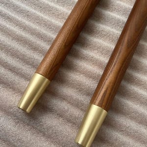 May include: Two wooden furniture legs with a warm brown finish and gold-colored metal tips. The legs are cylindrical and have a smooth, polished appearance, suggesting a mid-century modern design.
