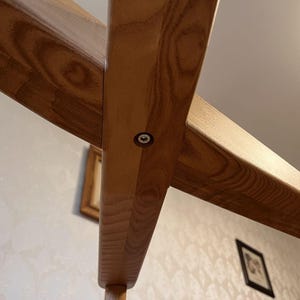 May include: Close-up of a wooden chair frame, showcasing the joinery. The light brown wood grain is visible, with a metal fastener at the intersection of two beams. The chair's design is simple and classic, with a focus on natural materials.