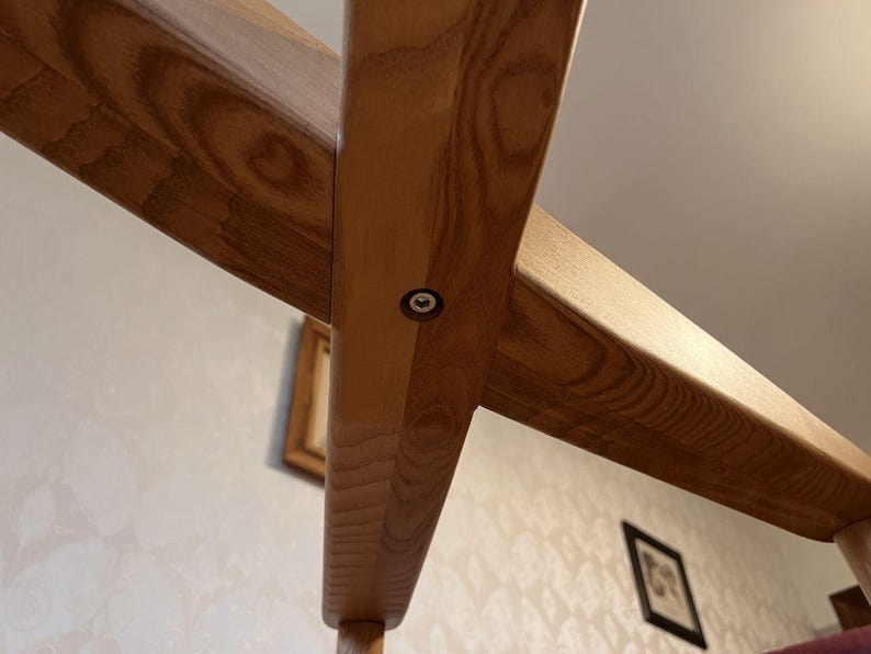 May include: Close-up of a wooden chair frame, showcasing the joinery. The light brown wood grain is visible, with a metal fastener at the intersection of two beams. The chair's design is simple and classic, with a focus on natural materials.