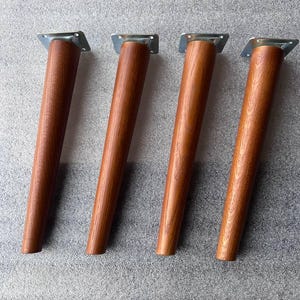 Può includere: Un set di quattro gambe per mobili affusolate con una finitura a venature del legno marrone caldo. Ogni gamba presenta una piastra di montaggio quadrata in metallo color argento sulla parte superiore con quattro fori preforati per il fissaggio.