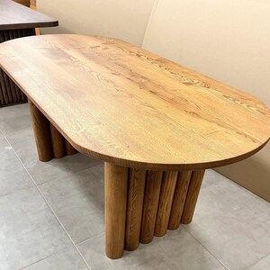 Puede incluir: Mesa de comedor ovalada de madera con un acabado marr&oacute;n claro. La mesa tiene una base &uacute;nica hecha de m&uacute;ltiples patas cil&iacute;ndricas de madera.