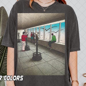 Op de afbeelding: Donkergrijs T-shirt met een grafische illustratie van een schilderachtig uitzicht met mensen en kijkmachines. Het kunstwerk toont een helderblauwe lucht met vogels die in de buurt van grote ramen vliegen. Het shirt heeft onderaan de tekst "R COLORS".