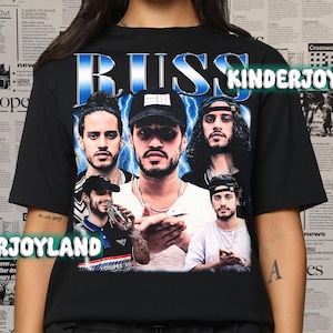 May include: Black t-shirt featuring a graphic of the musician Russ with the word "RUSS" in blue and white. The design includes multiple images of the artist. The word "KINDERJOY" is also visible.