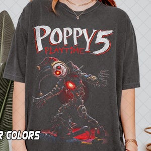 Puede incluir: Camiseta gris oscuro con el texto "POPPY 5 PLAYTIME" en blanco y rojo. La camiseta muestra un gráfico detallado de un personaje robótico con un sombrero de bufón y acentos rojos brillantes.