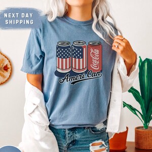 May include: Blue jean t-shirt featuring three soda cans with patriotic designs. Two cans display the American flag, while the third features the Coca-Cola logo. The phrase "Ameri Can" is written in cursive below the cans.