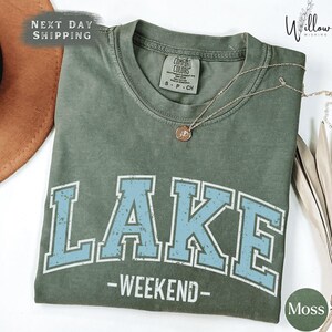 A green t-shirt with the word "LAKES" printed on it, along with the text "WEEKEND" below it. The t-shirt is displayed on a white surface, and there is a necklace visible in the background.