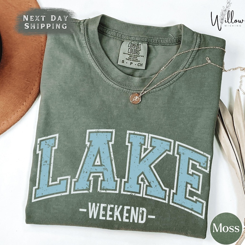 A green t-shirt with the word "LAKES" printed on it, along with the text "WEEKEND" below it. The t-shirt is displayed on a white surface, and there is a necklace visible in the background.
