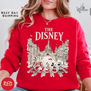 A woman wearing a red sweater with a graphic of Disney characters crossing a street.