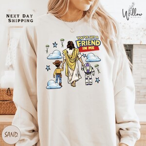 A person wearing a white sweatshirt with a graphic design depicting a cartoon-style illustration of a person holding a toy robot and a child, along with the text "You've got a FRIEND in me".