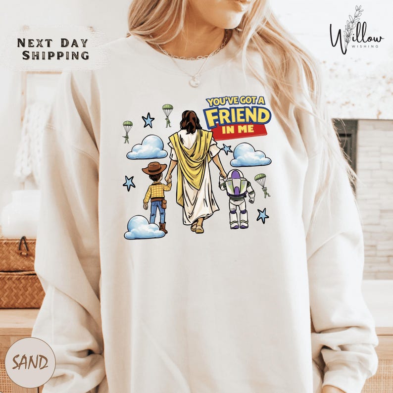 A person wearing a white sweatshirt with a graphic design depicting a cartoon-style illustration of a person holding a toy robot and a child, along with the text "You've got a FRIEND in me".