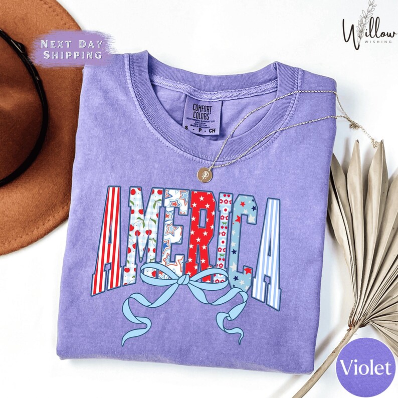 a purple t-shirt with the word "AMERICA" printed on it, along with a ribbon design. The t-shirt is laid out on a white surface, and there is a brown hat and a fan visible in the background.