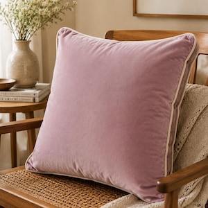 May include: A square throw pillow covered in soft, dusty rose velvet fabric. The pillow features a clean, piped edge detail in a contrasting light cream color that outlines the entire perimeter, providing a structured finish to the plush surface.