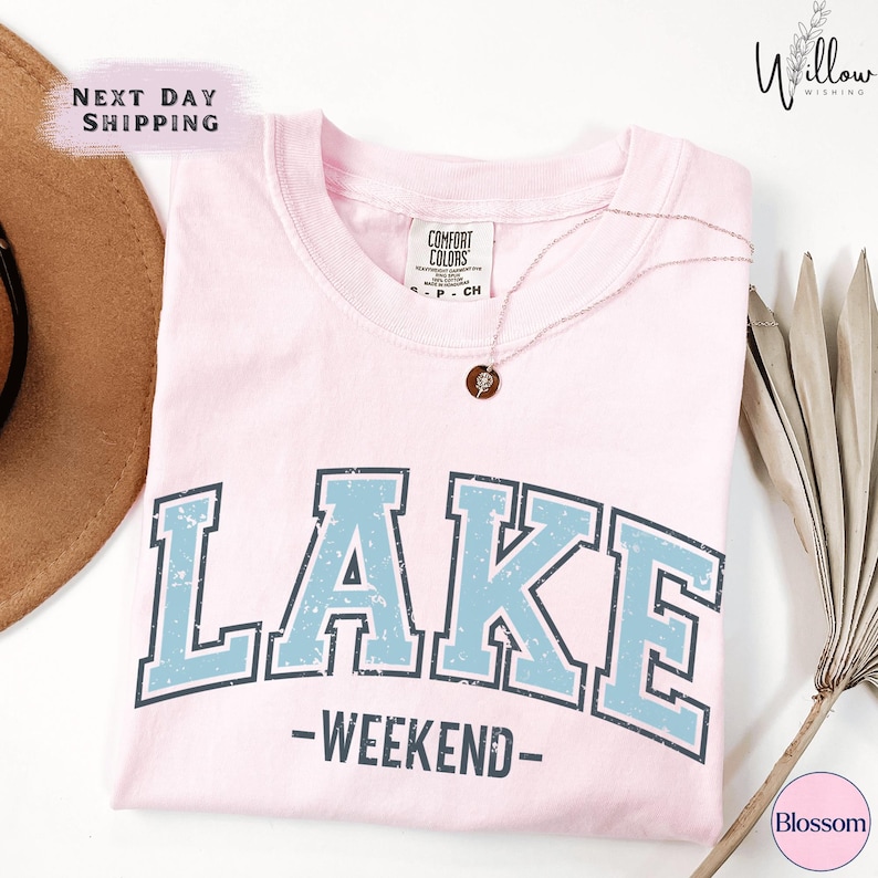 a pink t-shirt with the word "LAKES" printed on it, along with the text "WEEKEND" below it. The t-shirt is displayed on a white background, and there is a brown hat and some dried leaves nearby.