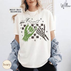 A woman wearing a white t-shirt with a graphic design featuring a bat, skeleton, and checkerboard pattern.