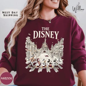 A woman wearing a maroon sweater with a graphic design featuring Disney characters crossing a street.