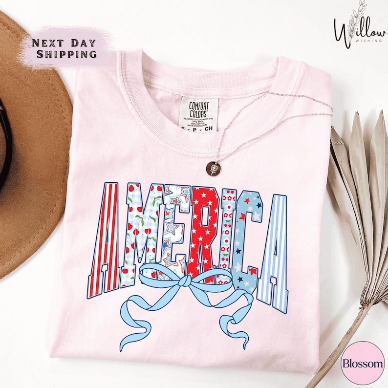 a pink t-shirt with the word "AMERICA" printed on it, along with a ribbon design. The t-shirt is placed on a white surface, and there is a hat and some leaves in the background.
