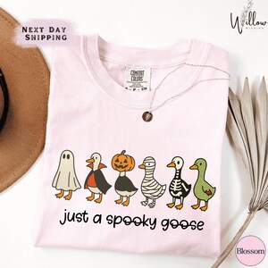 a pink t-shirt with a Halloween-themed graphic design, including cartoon-style illustrations of a ghost, skeleton, mummy, and a goose. The text on the t-shirt reads "Just a spooky goose", and there is a "Next Day Shipping" label above the t-shirt.