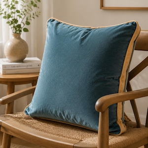 May include: A square throw pillow featuring a deep blue velvet fabric on the front and back, accented with a contrasting light tan or beige piping along the edges. The pillow has a soft, plush texture and a clean, simple design.