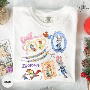 A white t-shirt with a colorful graphic design featuring various cartoon animal characters, including a rabbit, fox, and duck, along with decorative elements such as ribbons and bows.